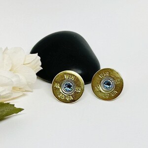 May include: A pair of gold-colored earrings made from shotgun shells with a blue gemstone in the center. The shells are marked "Win 20 Ga 44"