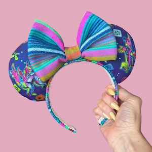 May include: A colourful headband with large ears and a vibrant striped bow. The ears are dark purple with a whimsical illustration. The headband is held by a hand with yellow nail polish.