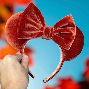 May include: A headband featuring two round ears covered in rust-colored velvet fabric. A large, matching rust-colored velvet bow is centered between the ears. The inner band is lined with a white, decorative trim featuring a repeating pattern.