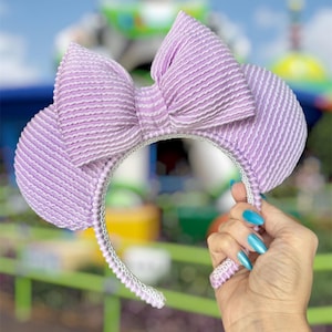 May include: A lavender headband featuring two round ears and a large, centered bow. The entire surface of the headband, ears, and bow is covered in a textured, seersucker-like fabric with thin, vertical white stripes throughout the design.