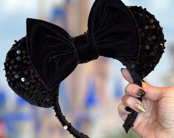 May include: Black sequined mouse ear headband with a large velvet bow. The ears are covered in black sequins, and the bow is made of soft velvet. The headband is held up against a blurred background of a castle.
