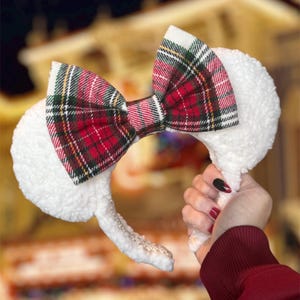 May include: White plush ear headband with a red, green, and white plaid bow. The headband has a silver-colored trim along the band. The ears are fluffy and soft. A festive accessory for themed events.