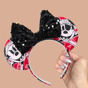 Peut inclure: Un serre-t&ecirc;te avec deux oreilles circulaires arborant un motif pirate rouge et noir avec une t&ecirc;te de mort. Un grand n&oelig;ud central noir recouvert de paillettes scintillantes est plac&eacute; entre les oreilles. L'arceau est bord&eacute; d'une garniture argent&eacute;e.
