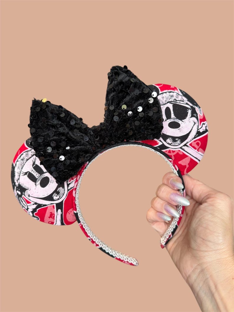Peut inclure: Un serre-t&ecirc;te avec deux oreilles circulaires arborant un motif pirate rouge et noir avec une t&ecirc;te de mort. Un grand n&oelig;ud central noir recouvert de paillettes scintillantes est plac&eacute; entre les oreilles. L'arceau est bord&eacute; d'une garniture argent&eacute;e.