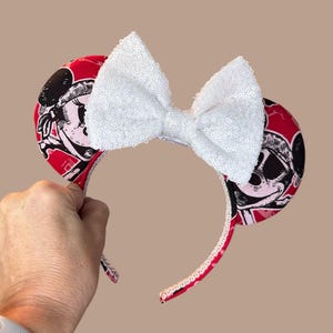 Peut inclure: Un serre-t&ecirc;te avec deux oreilles circulaires recouvertes d'un tissu rouge orn&eacute; d'un imprim&eacute; graphique noir et blanc repr&eacute;sentant un visage de personnage. Un grand n&oelig;ud blanc &agrave; paillettes est centr&eacute; entre les oreilles.