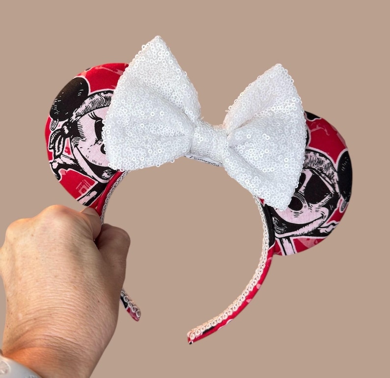 Peut inclure: Un serre-t&ecirc;te avec deux oreilles circulaires recouvertes d'un tissu rouge orn&eacute; d'un imprim&eacute; graphique noir et blanc repr&eacute;sentant un visage de personnage. Un grand n&oelig;ud blanc &agrave; paillettes est centr&eacute; entre les oreilles.