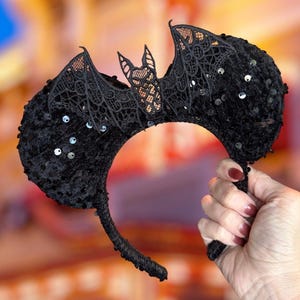 May include: Black Halloween headband with bat wings. The headband features a bat silhouette made of black lace and is embellished with sequins. The bat wings are detailed with intricate lace patterns, and the headband is held by a hand.