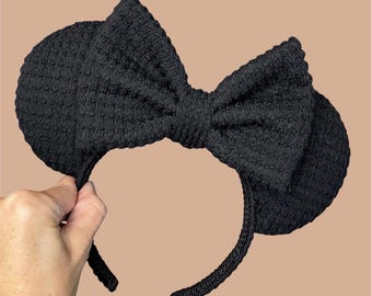 May include: A black headband featuring two circular ears and a large central bow, all constructed from a textured, waffle-knit fabric. The design is uniform in colour, with the fabric covering the entire headband, ears, and bow structure.