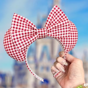 May include: Red and white gingham-patterned mouse ear headband. The ears are round and the bow is centered. The headband is held by a hand. The background is a blurred image of a castle and blue sky.