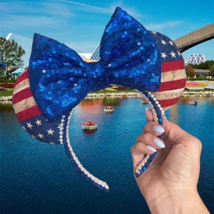 May include: A headband featuring two round ears decorated with a vintage-style American flag pattern of red stripes and white stars on a blue field. A large, bright blue sequined bow is centered between the ears, and the headband is lined with pearls.