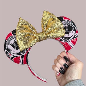 Peut inclure: Un serre-t&ecirc;te dot&eacute; de deux oreilles circulaires avec un imprim&eacute; graphique rouge, noir et blanc repr&eacute;sentant un personnage de dessin anim&eacute; portant un chapeau. Un grand n&oelig;ud central recouvert de paillettes dor&eacute;es scintille entre les oreilles.