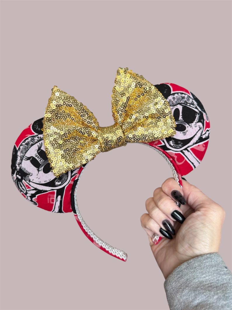Peut inclure: Un serre-t&ecirc;te dot&eacute; de deux oreilles circulaires avec un imprim&eacute; graphique rouge, noir et blanc repr&eacute;sentant un personnage de dessin anim&eacute; portant un chapeau. Un grand n&oelig;ud central recouvert de paillettes dor&eacute;es scintille entre les oreilles.