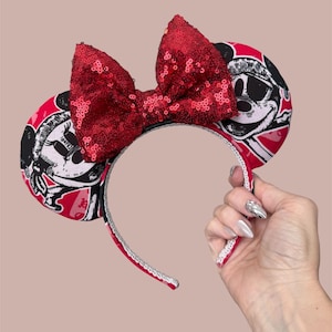 Peut inclure: Un serre-t&ecirc;te dot&eacute; de deux oreilles circulaires avec un imprim&eacute; graphique noir et blanc d'un personnage de dessin anim&eacute; sur fond rouge. Un grand n&oelig;ud central recouvert de paillettes rouges brillantes se trouve entre les oreilles.