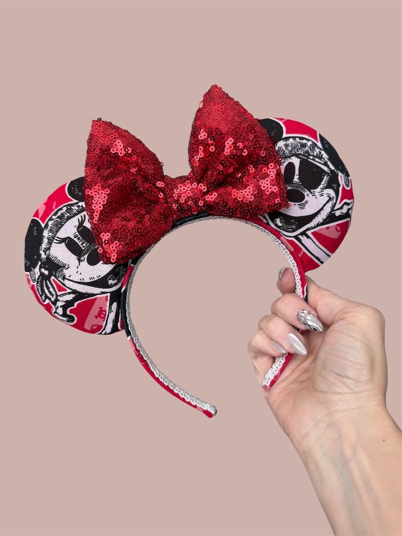 Peut inclure: Un serre-t&ecirc;te dot&eacute; de deux oreilles circulaires avec un imprim&eacute; graphique noir et blanc d'un personnage de dessin anim&eacute; sur fond rouge. Un grand n&oelig;ud central recouvert de paillettes rouges brillantes se trouve entre les oreilles.