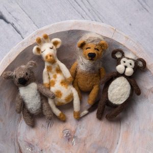May include: Four felt animal toys in a white bowl. The toys are a giraffe, a lion, a monkey, and a koala. The giraffe is brown and white, the lion is brown, the monkey is brown and white, and the koala is brown and white.