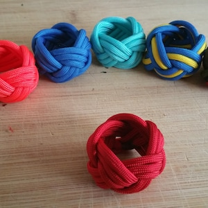 Könnte beinhalten: Sechs bunte Paracord-Armbänder, jedes mit einem anderen Knotenmuster. Die Armbänder sind aus rotem, blauem, grünem, gelbem und schwarzem Paracord.
