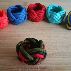 May include: Six colorful knotted rope rings, made with different colored paracord, on a wooden surface. The rings are in various colors, including red, blue, green, and yellow.