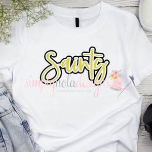 May include: White t-shirt featuring a gold and black "Saints" design. The design is outlined in black and filled with gold. The text "simplynoladesigns" is visible below the design.