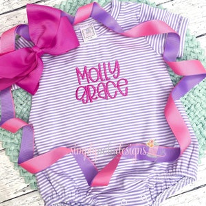 Pode incluir: Um body de bebé às riscas roxas claras e brancas, bordado com o nome "Molly Grace" em rosa. Um grande laço rosa e uma fita rosa e roxa estão dispostos à volta do body. O body tem mangas curtas e molas na parte inferior.