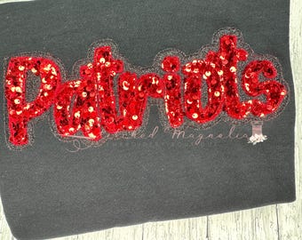 May include: Black fabric with the word "Patriots" in red sequins. The word is outlined in a silver-gray color. The text "Stitched Magnolia Embroidery Boutique" is below the word.