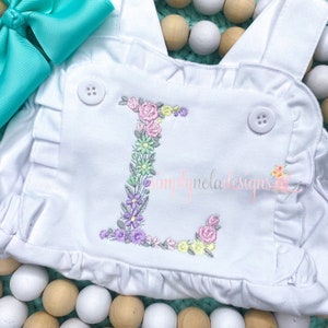 Pode incluir: Um macacão branco para bebê com uma letra L bordada com flores. O macacão tem um decote com babados e dois botões brancos. O macacão está sobre um fundo de contas de madeira.