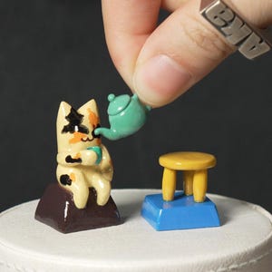 May include: A small, hand-painted figurine of a cat sitting on a brown rock, holding a teacup. A teal teapot is being poured into the teacup by a hand. The cat is sitting on a white cake stand with a blue and yellow miniature table and chair.