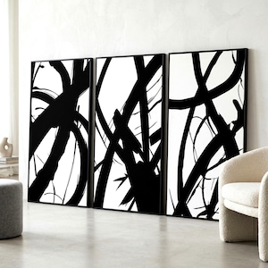 May include: Three framed abstract art pieces. Each piece features bold black brushstrokes against a white background. The artwork is displayed in a modern living space, with a neutral color palette and minimalist furniture.