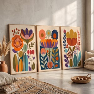 Könnte beinhalten: Drei gerahmte Kunstdrucke mit Blumenmotiven. Jeder Druck zeigt ein stilisiertes Blumendesign in leuchtenden Farben, darunter Orange, Lila und Blau, vor einem cremefarbenen Hintergrund. Die Rahmen sind hellbraun und das Kunstwerk wird an einer neutral getönten Wand ausgestellt.