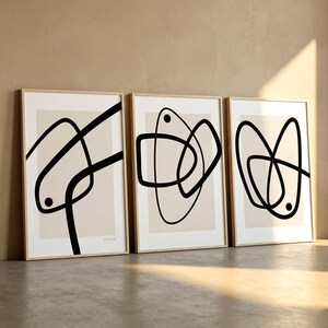 May include: Three framed abstract art prints. Each print features a beige background with black line art. The line art varies in shape and design, with some prints including small black circles. The frames are a light wood color.