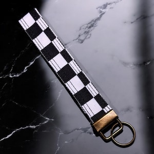 May include: A black and white checkered keychain with a gold metal key ring. The keychain is made of fabric and has a checkered pattern.