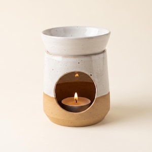 Könnte beinhalten: Ein Keramik-Wachswärmer mit zweifarbigem Design, bestehend aus einem gesprenkelten weißen Oberteil und einem beigefarbenen Sockel. Der zylindrische Körper hat eine kreisförmige Aussparung an der Vorderseite, in der eine brennende Teelichtkerze steht.