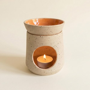 Op de afbeelding: Een gespikkelde keramische etherische olie diffuser met een ronde opening en een klein schaaltje. De binnenkant van het schaaltje en de opening zijn oranje. Er staat een brandende waxinelichtkaars in de diffuser, die een warme gloed creëert.