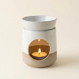 Op de afbeelding: Een keramische oliebrander met een witte en bruine buitenkant. De brander heeft een ronde opening met een brandende theelichtkaars erin. Bovenop staat een kleine, lichtgrijze kom.