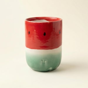 Può includere: Un bicchiere in ceramica con un design a forma di anguria. La metà superiore è rossa con dettagli di semi neri, il centro è bianco e il fondo è verde. Il bicchiere ha una forma cilindrica e una finitura liscia e lucida.