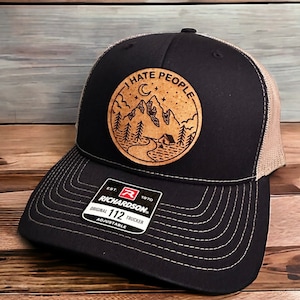 May include: A black and brown trucker hat with a brown leather patch that says "I Hate People" with a mountain scene and a crescent moon. The hat is adjustable and has a mesh back.