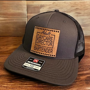 May include: A gray and black trucker hat with a brown leather patch that reads "Adult Daycare Director also known as Bartender". The hat is adjustable and has a Richardson 112 logo.