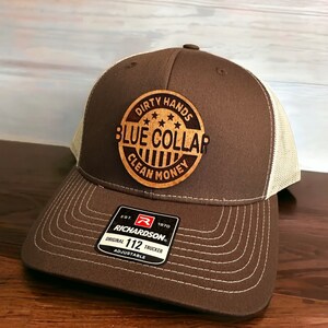 May include: Brown trucker hat with a brown leather patch that says "Dirty Hands Blue Collar Clean Money" with five stars and an American flag design. The hat is adjustable and has a mesh back.