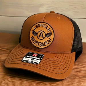 May include: A brown and black trucker hat with a leather patch that says "Asshole Merit Badge" with a winged letter A in the center. The hat is made by Richardson 112.