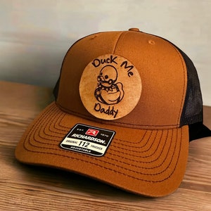 May include: Brown and black trucker hat with a brown leather patch that says "Duck Me Daddy". The hat is adjustable and has a Richardson 112 logo.