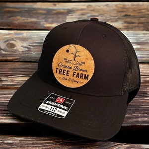 May include: Black trucker hat with a brown patch featuring the text "Charlie Brown Tree Farm - Cut & Carry" and the years "EST. 1965". The hat also has a red and white label with the text "RICHARDSON. ORIGINAL 112 TRUCKER ADJUSTABLE" and the year "EST. 1970".