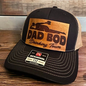 May include: A black and tan trucker hat with a brown leather patch that says "Dad Bod Drinking Team". The patch features a silhouette of a person lying down with a drink in their hand. The hat is adjustable and has a Richardson 112 logo.