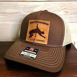 May include: Brown trucker hat with a brown leather patch that says "Mind your business, my wife is expensive". The patch features a silhouette of a person doing a pole dance. The hat has a white mesh back and a Richardson 112 label.
