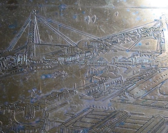 May include: A vintage brass plaque with an engraved image of a city skyline. The skyline features a bridge, buildings, and a stadium. The plaque has a worn, aged appearance.