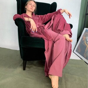 Puede incluir: Una mujer con una bata y pantalones de terciopelo rosa se sienta en un sill&oacute;n de terciopelo verde.
