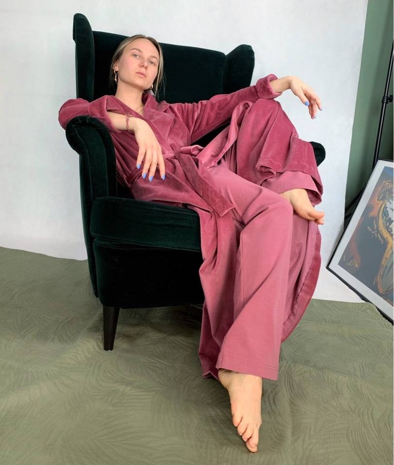 Puede incluir: Una mujer con una bata y pantalones de terciopelo rosa se sienta en un sill&oacute;n de terciopelo verde.