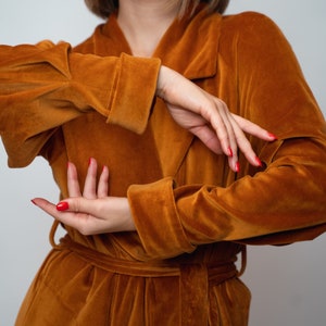 May include: A woman wearing a long, rust-colored velvet robe with a tie belt. The robe has long sleeves and a collar.