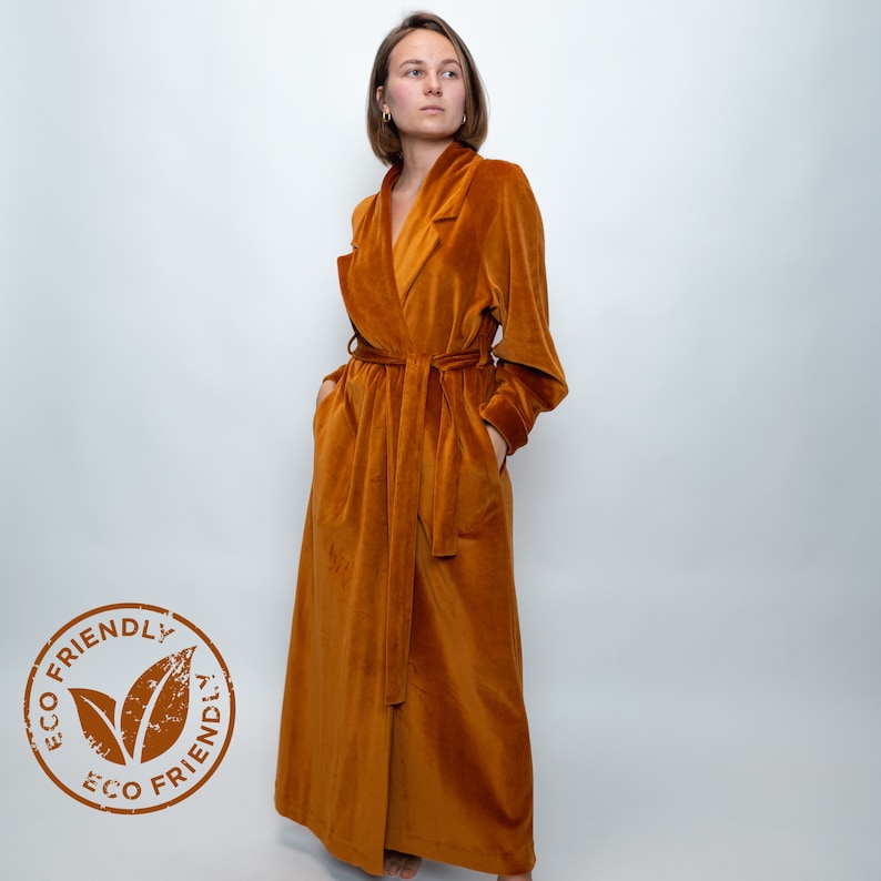 May include: A long, rust-colored velvet robe with a shawl collar and a tie belt. The robe has pockets on the sides. The robe is labeled "ECO FRIENDLY" with a green leaf logo.