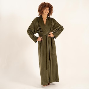 May include: An olive green, full-length robe with a shawl collar and a matching waist tie. The robe has long sleeves and pockets. The fabric appears to be a soft, plush material. The model is wearing a pearl necklace.