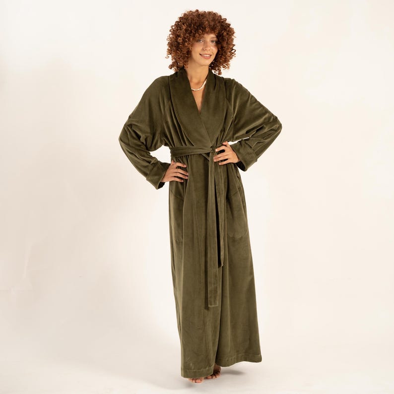 May include: An olive green, full-length robe with a shawl collar and a matching waist tie. The robe has long sleeves and pockets. The fabric appears to be a soft, plush material. The model is wearing a pearl necklace.