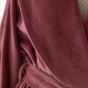 Puede incluir: Una bata de terciopelo rosa polvoriento con una cinta para atar. La bata tiene un cuello chal y mangas largas.